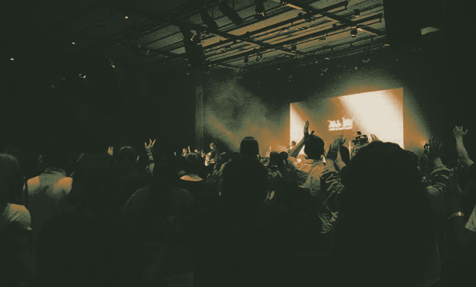 A room full of people with hands raised in worship, facing a lit stage.
